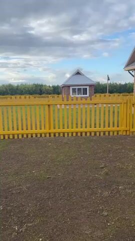 The video shows a panning shot of a rural landscape. In the foreground is a patch of tilled soil with some sparse green growth. A bright yellow wooden fence runs horizontally across the middle of the frame. Beyond the fence, there are two small wooden buildings. The building on the left is a small, square structure with a peaked roof and a window. The building on the right is a larger, log-style house with a grey metal roof, multiple white-framed windows, and decorative trim around the windows. The house has a visible chimney and a downspout. The background shows a line of trees and a cloudy sky. The scene appears to be an outdoor residential area, possibly a yard or a village.