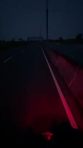 The video captures a first-person perspective from a moving vehicle, likely a motorcycle, at night or in the very early morning/late evening on a highway in Rupganj, Bangladesh.

The initial frame shows a bright red tail light and part of a hand at the bottom of the screen, indicating the viewpoint from the vehicle. The road is dark asphalt with clear white lane markings and a concrete barrier on the right side, which is illuminated by the red light.

As the video progresses, the camera pans up and slightly to the left, revealing an empty, multi-lane highway stretching into the distance under a dark blue-grey sky. Street lights on tall poles are visible further down the road. The surrounding landscape consists of low-lying vegetation and some distant, indistinct structures, possibly industrial or high-rise buildings. The overall scene conveys a quiet, solitary journey on a sparsely trafficked road during twilight or nighttime hours. No text is visible.
