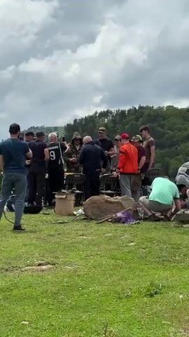 A group of men are gathered in a grassy area, with a forest and mountains in the background. Some men are standing around a fire pit, while others are talking. One man is wearing a black shirt with the number 9 on it. Another man is wearing an orange jacket. In the foreground, a woman is holding a phone and filming the scene. The weather appears to be cloudy.