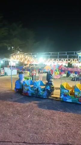 The video shows a nighttime scene at what appears to be a small amusement park or fairground. In the foreground, there is a children's train ride with several brightly colored carriages. The carriages are decorated with cartoon characters, including what looks like Batman on the front carriage, and other characters on the sides. The train is on a track and appears to be stationary. Behind the train, there are several stalls or tents, some decorated with string lights. People can be seen walking around and interacting in the background, though they are somewhat blurry due to the nighttime lighting and motion. The ground is covered in gravel or dirt. The overall impression is a lively but somewhat rustic nighttime entertainment venue.