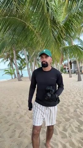 A man stands on a sandy beach, holding a camera. He is wearing a black long-sleeved shirt, striped shorts, a green baseball cap, and black gloves. Palm trees line the beach behind him. The ocean is visible in the distance. The man is looking towards the camera.