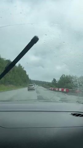 The video is filmed from inside a car driving on a wet road during a cloudy day. Water droplets are visible on the windshield, distorting the view of the road ahead. In the distance, several vehicles are driving, including a truck, a white van, and a few cars. The road is lined with trees on both sides, and there are some construction barriers in the right lane. The overall mood is somber and dreary due to the weather.