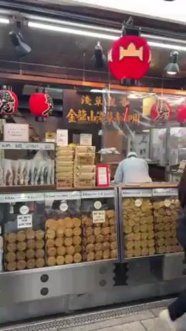 The video shows a crowded street in Japan. The street is lined with shops selling various goods, including food. There are many people walking along the street, including a man in a grey jacket and a black baseball cap. There are also traditional red lanterns hanging from the shops. The sky is dark, suggesting it is evening.
