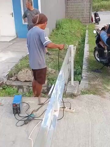 The video shows a man, wearing a blue shirt and brown shorts, welding a metal beam. He is standing next to a welding machine and holding a welding torch. The metal beam is long and appears to be part of a fence or railing. Another man, wearing a gray shirt and glasses, stands behind him watching. The scene takes place outdoors on a concrete surface, with grass and vegetation visible in the background. A motorcycle is parked to the right. The welding process generates sparks and bright light.