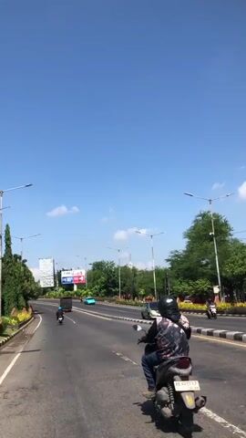 This is a vertical, first-person perspective video of a sunny day on a wide, multi-lane road in Buduran, Indonesia. The scene is dominated by a bright blue sky with a few scattered white clouds. The road is asphalt, with clear lane markings.

The primary subjects are several individuals riding motorcycles. In the foreground, a person wearing a dark hoodie and a black helmet is riding a light-colored scooter. Further ahead, other motorcyclists are visible, including one on a red scooter with a white shirt and red helmet, and others on darker scooters. A silver car is also seen driving in the same direction. The road is lined with trees and shrubbery, with decorative yellow and orange flowers along the roadside barriers. Streetlights are positioned at intervals along the road.

In the distance, a large billboard stands out, displaying advertisements. One advertisement has the text "10%" prominently displayed. Other structures and buildings are visible further back, interspersed with more trees. The overall impression is a bustling urban or suburban environment on a clear, bright day. The activity is the movement of traffic, with motorcycles and cars traveling along the highway. The time of day is clearly daytime, indicated by the strong sunlight and the blue sky. The weather appears to be warm and pleasant, typical of a tropical climate.