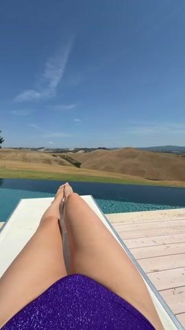 The video shows a first-person perspective of someone relaxing on a lounge chair by an infinity pool. The person is wearing a sparkly purple swimsuit bottom. Their legs and feet are visible, positioned towards the camera.

The setting is a scenic landscape with rolling, dry, tan hills under a clear blue sky with a few wispy clouds. An infinity pool stretches across the foreground, with its blue water contrasting with the dry landscape. The pool deck appears to be made of light-colored wood planks. A small tree is visible on the left side of the frame, near the edge of the pool. The scene suggests a warm, sunny day, likely during daytime. The overall impression is one of relaxation and enjoyment of a beautiful vacation spot. The location is identified as Asciano, Italy, which aligns with the Tuscan landscape depicted. No text is visible in the media.