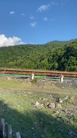 The video shows a panning shot of a natural landscape with a bridge. The scene is set in Yuxarı Tala, Azerbaijan, under a clear blue sky with a few wispy clouds. The time of day appears to be morning or late afternoon, indicated by the bright, soft sunlight.

The foreground is a rocky, uneven terrain covered with sparse green grass and scattered stones. In the middle ground, a multi-span bridge with an orange and red superstructure is visible. The bridge is supported by concrete pillars and appears to be crossing a dry riverbed or a shallow body of water. There are also some pipes running alongside the bridge.

The background is dominated by a lush, green, tree-covered mountainside, indicating a forested area. The dense foliage suggests a healthy and vibrant natural environment. The overall atmosphere of the video is peaceful and serene, highlighting the beauty of the natural landscape. There is no visible text in the video. The camera pans from left to right, revealing more of the bridge and the surrounding mountainous terrain.