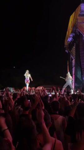A night concert is captured from the perspective of the audience. On stage, a female singer in a shimmering outfit stands center stage, singing into a microphone. To her right, a guitarist plays an electric guitar. The stage is brightly lit, with spotlights illuminating the performers. In front of the stage, a large crowd of people is visible, many with their hands raised in the air, some holding up phones to record. The overall atmosphere is energetic and lively, with the dark night sky serving as a backdrop.