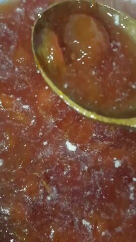 The video shows a close-up of a wooden spoon scooping a thick, reddish-orange jam from a pot. The jam appears to be homemade, with visible chunks of fruit and a slightly cloudy texture. Small white flecks, possibly foam or impurities, are scattered throughout the jam. The lighting suggests an indoor setting, likely a kitchen, and the time of day is not discernible. The focus is on the texture and movement of the jam, giving a sense of its consistency. There is no visible text. The scene evokes a feeling of domesticity and the preparation of food. The location context of Smarhon, Belarus, suggests this could be a traditional fruit preserve being made.