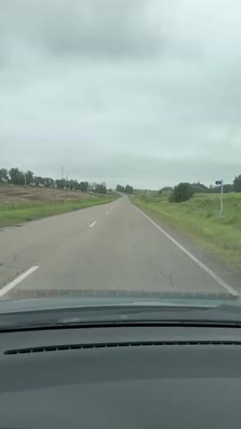 The video shows a first-person perspective from inside a vehicle traveling on a two-lane road. The road is asphalt and appears to be in fair condition with some cracks. The vehicle is moving forward, and the view is of the road ahead and the surrounding landscape. The landscape consists of rolling green hills with patches of trees and some agricultural fields on either side of the road. The sky is overcast and gray, suggesting cloudy weather. There are no other vehicles or people visible in the immediate vicinity. The dashboard of the vehicle is visible at the bottom of the frame. The overall scene depicts a drive through a rural or semi-rural area under a cloudy sky.