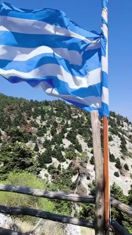 A Greek flag waves in the wind in front of a mountainous landscape under a clear blue sky. The flag is positioned on the right side of the frame, attached to a wooden pole. The pole is supported by a rustic wooden railing in the foreground. The mountains in the background are covered with dense green trees and rocky terrain. The camera is panning slightly to the right, capturing the movement of the flag and the expanse of the landscape.