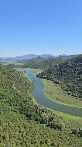 This is a vertical, high-angle drone shot overlooking a winding river through a lush, green, mountainous landscape. The river is wide and bends to the right, bordered by dense vegetation and reeds. The surrounding hills are covered in trees and greenery, with a clear blue sky above. The video pans slowly across the landscape, showcasing the natural beauty of the area. There are no people or man-made objects visible in the frame.