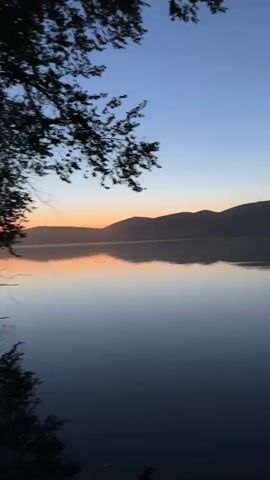 This media file captures a serene, panoramic view of a tranquil lake at dawn in Milyanfan, Kyrgyzstan. The scene is dominated by the calm water, which beautifully mirrors the dusky sky and the silhouetted landscape.

The primary subject is the natural scenery. In the foreground, the dark, still surface of the lake reflects the sky and distant hills. The water appears calm, with only subtle ripples breaking the perfect reflection. As the eye moves towards the horizon, a range of gently sloping hills is visible, their shapes softened by the low light. Above the hills, the sky transitions from a pale orange and pink glow near the horizon, indicating the very early stages of sunrise, to a deep, clear blue overhead. A few tree branches, silhouetted against the sky, are visible in the upper left corner of the frame, adding a touch of foreground detail.

The activity taking place is simply the passage of time, as the day transitions from night to morning. There are no people or man-made objects clearly visible, emphasizing the natural beauty and peacefulness of the location. The overall mood is one of tranquility, quietude, and the awe-inspiring beauty of nature at the start of a new day. The time of day is clearly early morning, with the sun just beginning to rise. The weather appears calm and clear, conducive to the still water and unobstructed view of the horizon. The location is identified as Milyanfan, Kyrgyzstan, suggesting a remote, natural setting. There is no visible text in the media file.