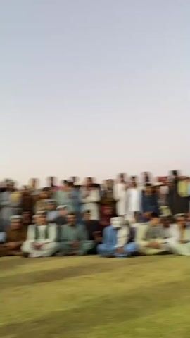The video shows a group of men and boys gathered outdoors on a grassy field. The sky is a pale, clear color, suggesting it is either early morning or late afternoon. Many of the people are dressed in traditional Islamic clothing. Some men are seated on the ground in the foreground, while others stand or are partially visible in the background. One man in the center is wearing a white Guy Fawkes-style mask. There are several individuals in the video who appear to be security personnel, including one man in tactical gear holding a rifle. The latter part of the video shows a man in a green and white striped soccer uniform receiving an orange box from another man who is wearing traditional clothing. Other men stand around them, some observing and one seemingly speaking. The overall atmosphere appears to be a community gathering or event, possibly a sports award ceremony.