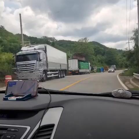 This is a first-person perspective video from inside a car. The car is stopped on a road, likely due to traffic congestion. In the foreground, a smartphone is mounted on the dashboard, displaying a navigation app. The road ahead is visible, with several large trucks parked or moving slowly. One truck is laden with green cylindrical objects, possibly crates of produce. Another truck is a flatbed carrying what appears to be construction materials. There are also forklifts and workers visible near the trucks, suggesting a loading or unloading operation. The environment is rural, with trees and hills in the background under a cloudy sky. The time displayed on the car's stereo is 1:32.