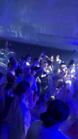 A crowded indoor party is depicted, with a diverse group of people dancing and socializing under blue and white lighting. Smoke machines create a hazy atmosphere, and the ceiling appears to be draped with fabric, possibly for decoration or to diffuse the lighting. Several individuals are holding their phones up, likely recording or taking pictures of the event. The overall ambiance suggests a celebratory or festive occasion.