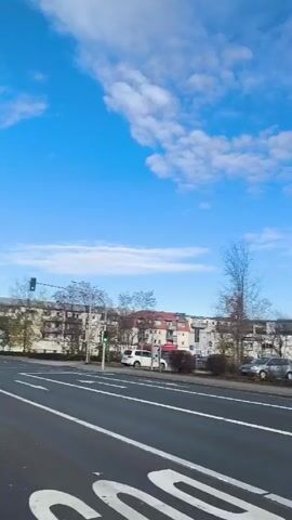 The video is a point-of-view shot taken from a car moving along a road in a town on a bright, partly cloudy day. The road is paved with white dividing lines. On the right side of the road, there are parked cars, vans, bushes, and buildings of varying heights. The sky is blue with puffy white clouds. The car moves past a street light, a parking lot, and then turns right at a traffic light. A hill is visible in the background. Other cars are also on the road.