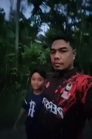 The video shows a man and a boy standing next to each other, looking at the camera. The man is on the right, wearing a black polo shirt with red and white designs and the word "PERSIJAP" printed on it. He has short dark hair and a mustache. The boy is on the left, wearing a dark blue t-shirt with a white letter "A" on it. He has short dark hair and is smiling slightly. They appear to be standing on a path or road, with greenery and trees in the background. The lighting suggests it is either dusk or dawn. The camera is shaky, suggesting it is handheld.