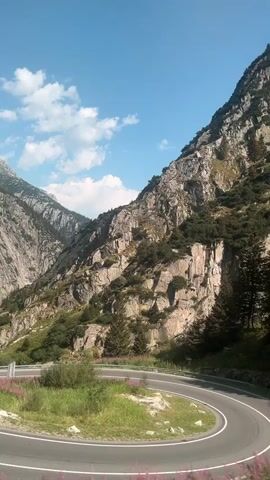The video shows a winding mountain road with steep, rocky cliffs on one side and trees on the other. The road is paved and has white lines marking the lanes. There are some small purple flowers growing alongside the road. The sky is blue with a few white clouds. The video appears to be taken from a moving vehicle, as the scenery is slightly blurred.