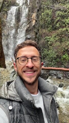 A man with brown hair, glasses, and a gray vest smiles at the camera in a selfie. Behind him is a waterfall cascading over rocks into a stream. Trees surround the waterfall, and a wooden railing is partially visible. The man makes a fist pump. The background is slightly blurred, emphasizing the man in the foreground.