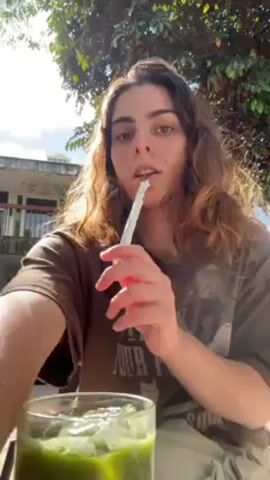 A person with long, wavy brown hair and wearing a brown t-shirt is sitting outside, stirring a green drink with ice in a glass. The person holds a straw in their mouth for a moment, then switches to a brown straw to stir the drink. The background includes a building and green trees, with sunlight filtering through the leaves. The person's left hand has visible tattoos.