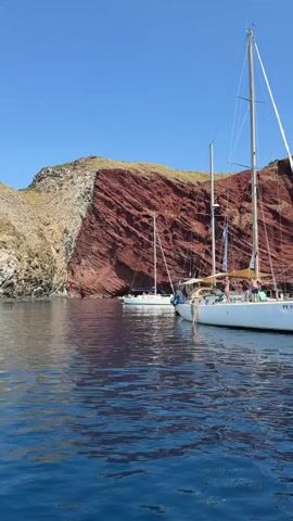 The video shows a scenic view of two sailboats anchored in clear blue water near a large, reddish-brown cliff face. The sailboat in the foreground is larger and white, with its sails furled. A person can be seen on its deck, leaning over the side. The second sailboat is smaller and white, anchored further in the distance, also with its sails furled. The cliff face is steep and layered, with patches of vegetation on its upper slopes. The sky is clear and blue, and the water is calm with ripples reflecting the sunlight and the sailboats. The overall scene is peaceful and suggests a leisurely day on the water in a beautiful natural setting.
