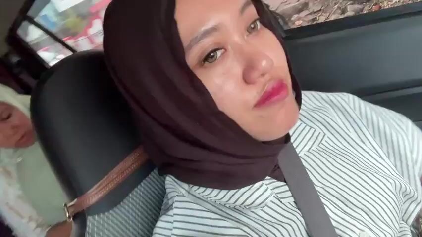 The media file is a close-up selfie video of a young woman wearing a dark brown hijab and a white and grey striped shirt. She is seated in the passenger seat of a car, with the camera tilted at an angle. A seatbelt is visible across her chest. Her makeup includes bright pink lipstick and some eye makeup. She smiles and laughs directly at the camera, showing off her braces in one of the frames. In the background, out of focus, a child can be seen in the back seat of the car. The view through the car window suggests an outdoor setting, possibly a street with some vegetation and buildings. The lighting appears to be natural daylight, suggesting it is daytime. The overall mood is cheerful and lighthearted. There is no visible text in the media file. The context suggests the location is Sumbersari, Indonesia.