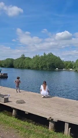 The video shows a wide shot of a serene outdoor scene. In the foreground, a wooden pier extends into a calm body of blue water, likely a river or lake. On the pier, a young child, dressed in a grey top and patterned trousers, stands near the edge, facing the water. To their right, a woman with long brown hair, wearing a white top and light-colored trousers, is crouched at the very edge of the pier, also looking down at the water.

The background features lush green trees lining the far bank of the water. Beyond the trees, the sky is a vibrant blue with scattered white, fluffy clouds. On the distant bank, a few small white objects, possibly boats or structures, are visible. The overall ambiance is peaceful and calm, suggesting a pleasant day spent outdoors. The time of day is clearly daytime, with bright sunlight illuminating the scene. The location, Kipen, Russia, implies a natural or recreational setting. There is no visible text in the video.