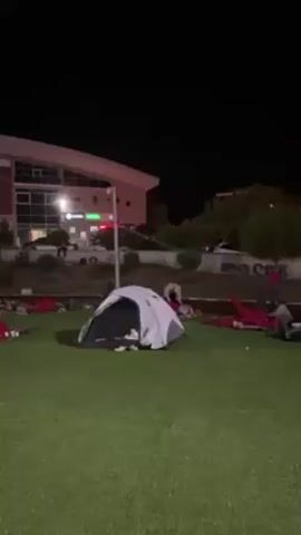 The video depicts a nighttime scene on a patch of artificial turf in front of a large building with a curved roof. Several people are visible either lying on the grass, wrapped in red blankets, or standing near the building. A single small tent stands on the turf. The area is dimly lit, and the overall atmosphere appears to be calm and quiet.