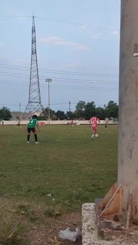 The video shows an outdoor soccer game with several players in uniforms on a grassy field. There is a tall transmission tower in the background, along with some lights and power lines. In the foreground, a person stands partially in view, to the right of the frame, obscuring part of the field. The sky is visible in the background with some clouds. The players are scattered across the field, some kicking or dribbling a ball. The action appears to be in progress.