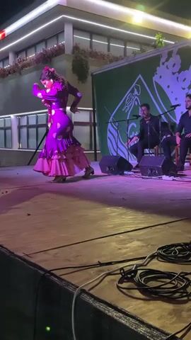 A woman in a purple polka-dot flamenco dress with ruffles dances on a wooden stage. She has her hair pulled up and is wearing earrings. Two men are seated behind her on the stage. One man plays a stringed instrument, possibly a guitar or violin, and the other appears to be singing into a microphone. A green banner with a white logo is hung behind the musicians. The stage is lit with purple and yellow lights, and the background shows a building with lit windows and exterior lights. Cables are visible in the foreground.