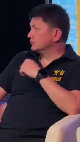 A medium shot shows a seated man in profile facing left. He has dark hair and is wearing a navy polo shirt with a yellow and gold design on the left chest. The design appears to be a trident shape with a stylized figure above the words “MYKOLAIV REGION”. He also wears a black wristwatch with a silver face. He has his hands clasped in his lap. The background is a screen with indistinct colors. The man appears to be listening intently. The seat on which he is sitting is white.