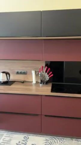 The video shows a modern, open-concept kitchen and living area. The kitchen features two-tone cabinetry, with dark gray upper cabinets and a burgundy color on the lower cabinets. The countertops are a light wood grain. Appliances such as a kettle, knife block, cooktop, toaster, and coffee maker are visible. The sink and faucet are black. A dishwasher door is open, revealing the interior. The living area includes a round, dark-colored dining table with red chairs. A patterned rug is under the table. A TV and a cabinet with drawers are against the wall near a sliding glass door, which is covered with sheer and patterned curtains. An air conditioner unit is mounted on the wall above the door. The room has pale yellow walls and wood-look flooring. There are no people visible in the video.