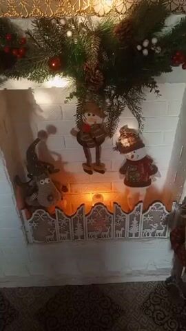The media file depicts a Christmas-themed mantelpiece. The mantelpiece is decorated with a garland of evergreen branches, red berries, pinecones, and white baubles. Several plush holiday figures are arranged on the mantel, including a reindeer with antlers and a hat, a figure resembling a snowman wearing a scarf and hat, and another snowman dressed in a festive sweater and hat. The background is a white brick wall, and a simulated fireplace with glowing orange and yellow light is visible beneath the figures. A decorative screen with cutouts of trees and deer separates the fireplace from the viewer. The overall impression is one of a cozy and festive holiday decoration, likely set in a home environment in Petukhovo, Russia, though no specific time of day or weather conditions can be determined. There is no visible text in the media file.