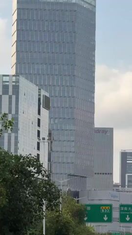 The video shows traffic moving along a city street lined with trees. Several tall buildings are visible in the background, including one under construction. There are green road signs with arrows and Chinese characters, indicating directions to different areas like Shennan Blvd and Baishi Rd. The traffic includes cars, taxis, and a small truck. The sky is overcast, but the scene is generally well-lit. The view is from a slightly elevated position, likely from a vehicle moving with the traffic.