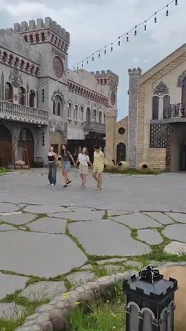 The video shows four young women holding hands and dancing in a courtyard in front of a stone building with turrets. The building appears to be a castle or a similarly styled structure. The ground is paved with large, irregular stones. String lights are visible above the courtyard. The sky is overcast. The women are dressed casually. Their movements are synchronized as they dance.