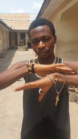 A young, light-skinned man stands outdoors, facing the camera, wearing a black tank top with a white trim and a small white logo on the front, several beaded bracelets on his left wrist, and multiple gold and silver-colored bracelets on his right wrist. He is also wearing a large, gold-colored necklace. The background shows what appears to be a narrow alleyway between two buildings, with a corrugated metal roof visible above. He is moving his hands up to his face, and then down, as if performing a brief, self-conscious gesture. The overall lighting suggests a sunny day.