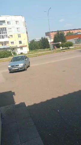 The video shows a daytime scene on a street in Polysayevo, Russia. The sun is shining, indicating good weather. In the foreground, a grey sedan is driving towards the viewer on a paved road. The car has a license plate that reads "C892CC 77". Behind the car, on the left side of the frame, is a multi-story apartment building with a light-colored facade and blue window frames. There are trees and other buildings visible in the background, suggesting an urban or suburban environment. A street lamp is also present. On the right side of the road, in the distance, there are more trees and a building with a red roof. A person is walking on the sidewalk in the background. The overall impression is a typical street view on a clear day. There is no discernible emotional tone or particular event occurring beyond the normal flow of traffic. The visible text is limited to the license plate of the car.