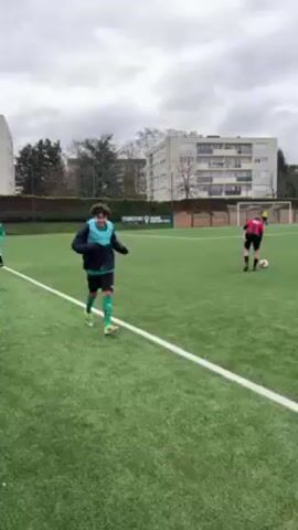 The video shows a soccer field with a few people on it. A young man is running toward the camera, smiling. He is wearing a black jacket, a light blue training bib, black shorts, and green socks and cleats. In the background, there are two other players and a goal post. The field is green and surrounded by a fence and some buildings. The sky is overcast.