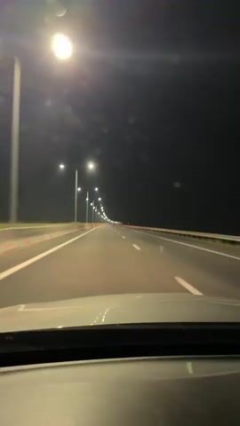 The video shows a point-of-view shot from inside a vehicle traveling on a highway at night. The highway is illuminated by streetlights that are spaced along the side of the road. The road itself is paved and has clear lane markings. On the left side of the road, there are concrete barriers with red and white reflectors. The sky is dark, indicating it is nighttime. The overall impression is one of a smooth, continuous journey through a dark, well-lit, and empty highway. The location is identified as Chibat'is Temi, Georgia. There are no visible people or other vehicles in the immediate vicinity, suggesting a solitary drive. The mood is calm and peaceful, with the steady movement of the car and the repetitive pattern of streetlights creating a hypnotic effect.