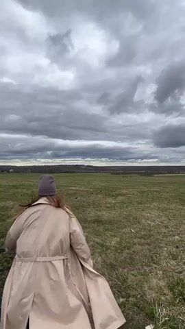 A person wearing a beige coat and a dark beanie is running away from the camera across a large, grassy field on a cloudy day. The field has patches of green and brown grass, suggesting the transition of seasons. In the distant background is a line of trees and buildings, indicating a town or city. The sky is filled with heavy, grey clouds.