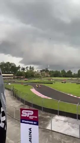 A video shot from an elevated angle, shows a go-kart race on a small track. The race is taking place on a cloudy day. The track is surrounded by green grass and trees in the background. There are multiple go-karts racing, mostly red. The video shows a sign for Scooter Prix sponsored by Yamina. There is a black flag visible on the left side of the screen, partially obstructing the view. A plane can be seen flying in the sky in the background.