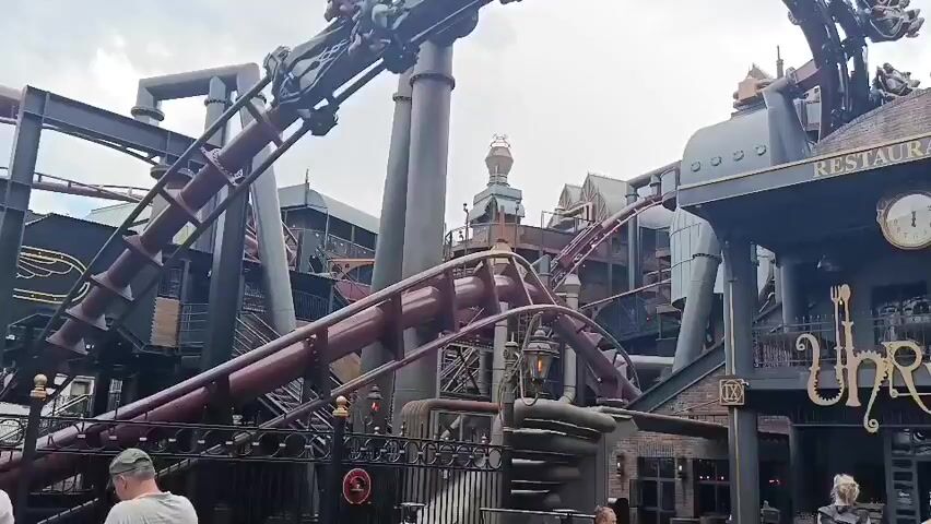 A roller coaster train with riders is visible on a steep, curved track. The train is dark with accents of red and gold. The track is also dark red and supported by large, dark grey structures. In the background, there are buildings with a steampunk aesthetic, featuring pipes, gears, and intricate designs. The sky is cloudy. In the foreground, two people are standing near a black metal fence, looking towards the roller coaster. The person on the left is wearing a white top, and the person on the right is wearing a grey cap and a light-colored shirt.