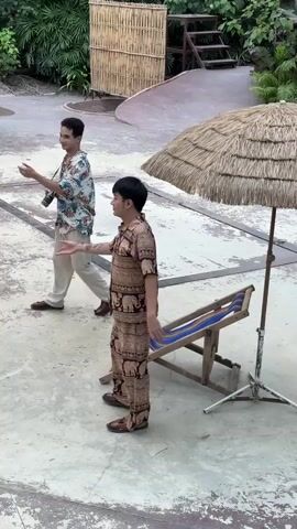 The video opens with two men standing on a concrete patio. One man, wearing a white shirt with a floral pattern and tan pants, stands facing left with his arms outstretched. The other man, wearing a matching tan outfit with an elephant pattern, stands facing right, also with his arms out. Behind them, a thatched umbrella shades a wooden lounge chair. In the background, there is bamboo fencing and lush greenery.

At 00:01, a woman wearing a flowing purple dress and a large purple hat enters the frame from the left, approaching the man in the floral shirt. She holds her hands out towards him.

By 00:02, the woman has taken the man's hands and is turning towards the lounge chair. The man in the elephant pattern outfit remains facing right.

At 00:03, the woman is seated in the lounge chair, still holding the hand of the man in the floral shirt. The man in the elephant pattern outfit is now facing the other two and gesturing with his hands.

By 00:04, the woman is fully settled in the chair, and the man in the floral shirt is leaning towards her, still holding her hand. The man in the elephant pattern outfit is standing between them, looking at them. The scene appears to be set outdoors, possibly in a garden or resort area.
