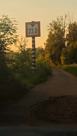 The video shows a road sign indicating a height restriction of 23.0. The sign is on a striped pole. The road appears to be unpaved and somewhat damaged. The surroundings are green and lush with trees and vegetation. The sky is a warm orange and yellow, suggesting it is either sunrise or sunset. The camera is moving forward along the road.