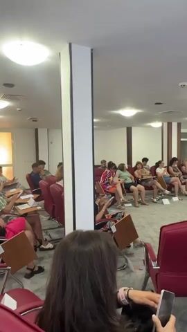 The video shows a group of people seated in a room that appears to be a conference or seminar space. The room has a carpeted floor, a white ceiling with several lights, and walls with decorative wood paneling. There are rows of red chairs, some occupied by individuals, facing a stage area at the front of the room. On the stage, there is a projector screen displaying an image, a table with a flip chart, and a podium. Two people are standing near the podium and projector screen, presumably the speakers or presenters. One person is a man in a light-colored shirt and pants, and the other is a woman in a dark dress. The audience members are mostly women, dressed casually, and appear to be listening to the presentation. Some individuals in the foreground are holding phones or papers. The overall atmosphere suggests a formal gathering or educational event.