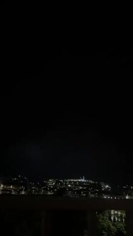 The video shows a night view of a city with distant lights, captured from an elevated position.  In the distance, a bright object appears to be a tall building or structure illuminated against the dark sky.  A burst of bright lightning illuminates the sky above the city. The foreground shows what appears to be a railing.  Some foliage is visible below the railing.