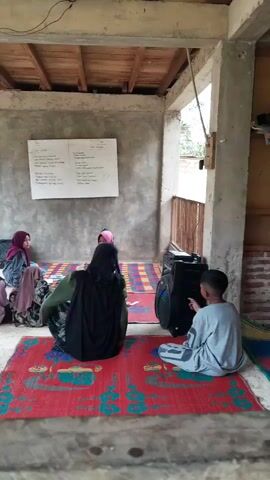 The video shows a group of young people sitting on prayer rugs in a simple, open-air structure. Five young girls, wearing hijabs in various colors, are gathered in a semicircle facing a woman also wearing a hijab. On the right side of the frame, a young boy in a gray outfit sits cross-legged next to a portable speaker. A whiteboard with text is visible on the wall behind them. The structure has exposed wooden rafters, concrete walls, and a partial fence on the right side. The atmosphere appears to be one of learning or group activity.
