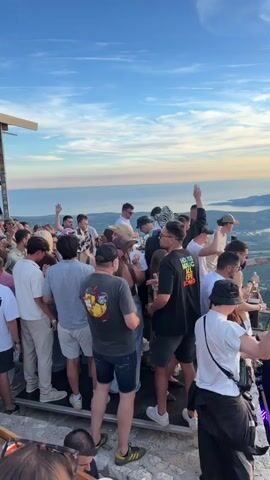 A crowded outdoor gathering is happening on a raised platform, possibly a rooftop or viewpoint, with a panoramic view of a coastal landscape and mountains under a partly cloudy sky. A diverse group of people, predominantly young adults, are standing and mingling, many holding drinks. The atmosphere appears festive, with some individuals dancing or gesturing enthusiastically. Several large speakers are visible, suggesting music is being played, possibly a DJ set. The time of day appears to be late afternoon or early evening, indicated by the warm, golden light illuminating the sky and the distant scenery. The people are dressed in casual summer attire.