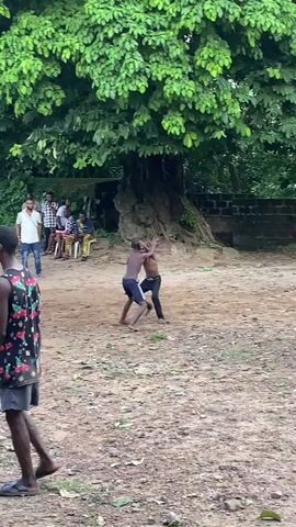 In a video recorded in Unwana, Nigeria, two young men are seen wrestling on a dusty, open ground. They are shirtless, wearing dark shorts, and are actively grappling with each other, attempting to take their opponent down.

The wrestling match takes place outdoors, under the shade of a large, mature tree with dense green foliage. Several spectators are gathered around the wrestling area. Some men are standing, casually dressed in t-shirts and jeans, while others are seated on chairs or benches further back, also under the tree's shade, observing the match intently.

The ground is dry and earthen, with scattered leaves and a discarded plastic bottle visible in the foreground. The overall atmosphere suggests a community gathering or a traditional sporting event, likely occurring during the daytime. One spectator in the immediate foreground, partially visible, is wearing a patterned shirt and flip-flops. The scene depicts a lively local event with engaged onlookers.